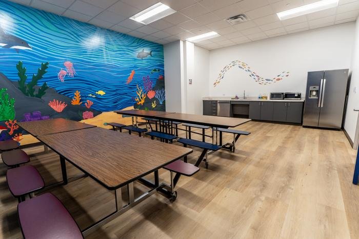 The lunch room at SOS McKinney: folding cafeteria tables in front of a painted underwater mural, with a kitchen counter and refrigerator along the far wall.